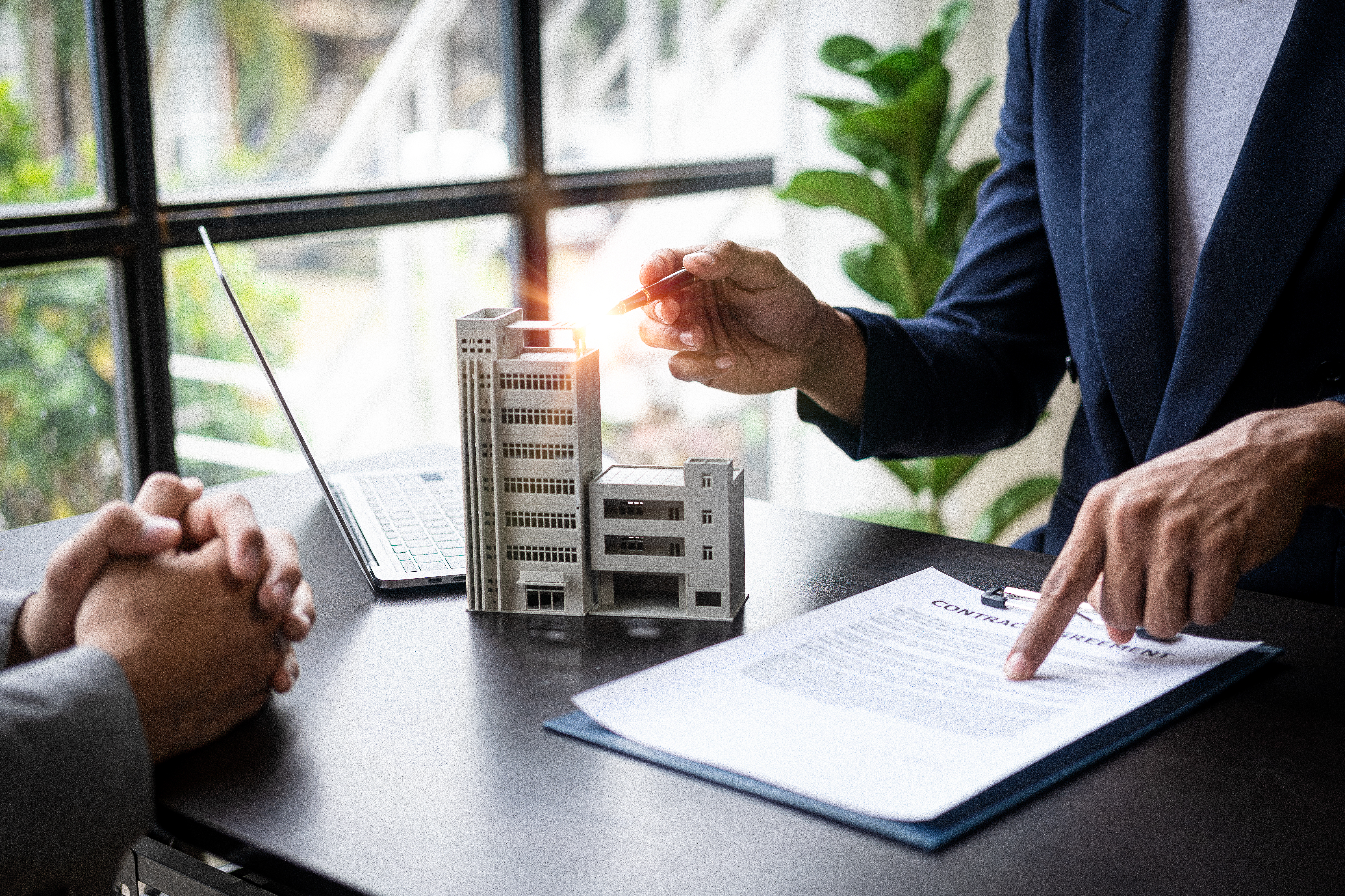 A figurine of a tall building next to a person passing another person a pen while pointing to a document